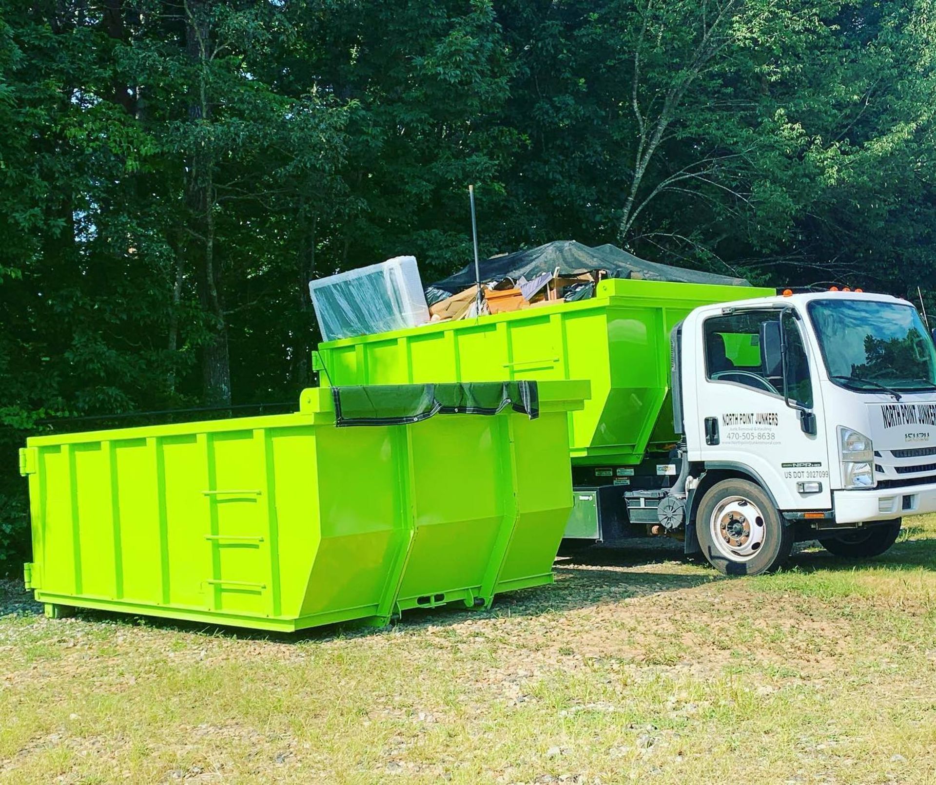 A green dumpster is sitting next to a white truck in a field.