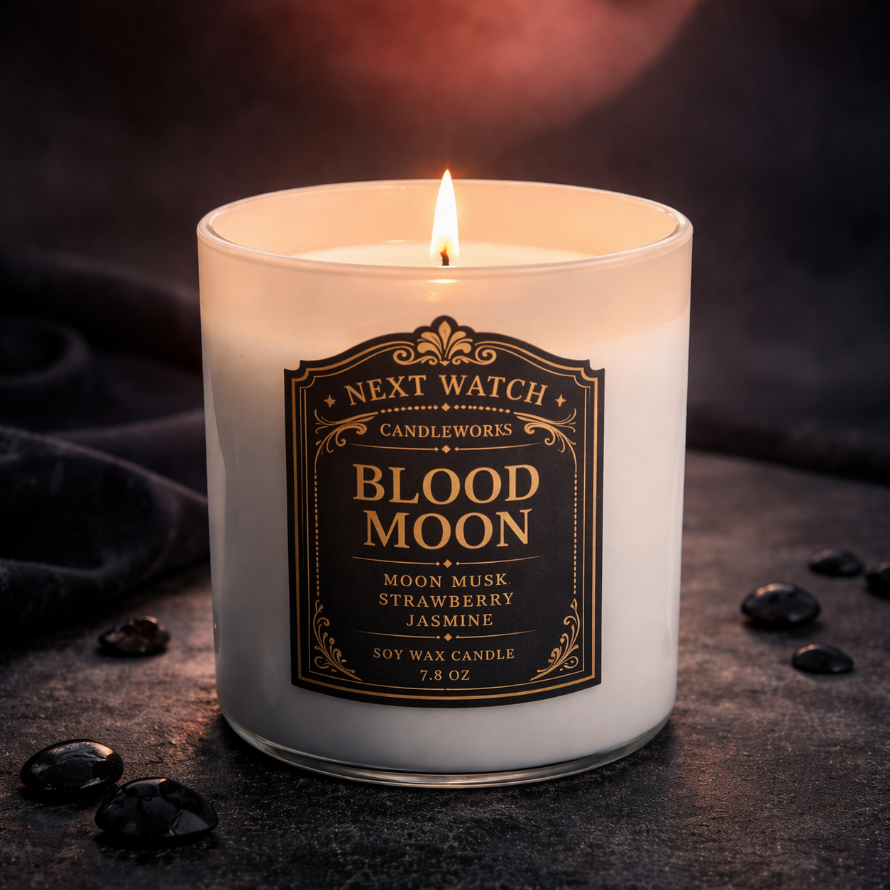 Lit white candle labeled “Blood Moon” with a black-and-gold label, surrounded by dark stones.