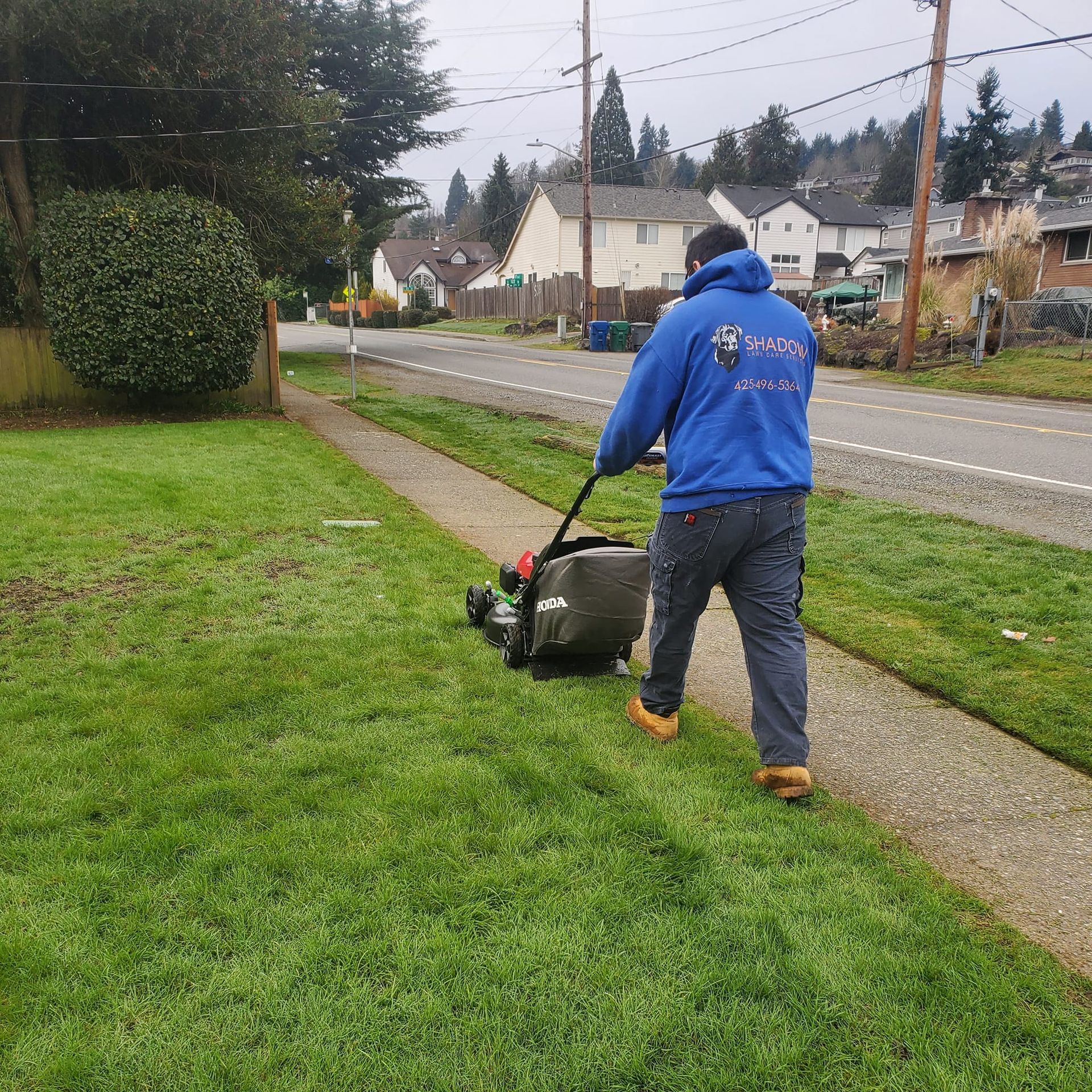 A person in blue hoodie mows a lawn next to a sidewalk and street on an overcast day.