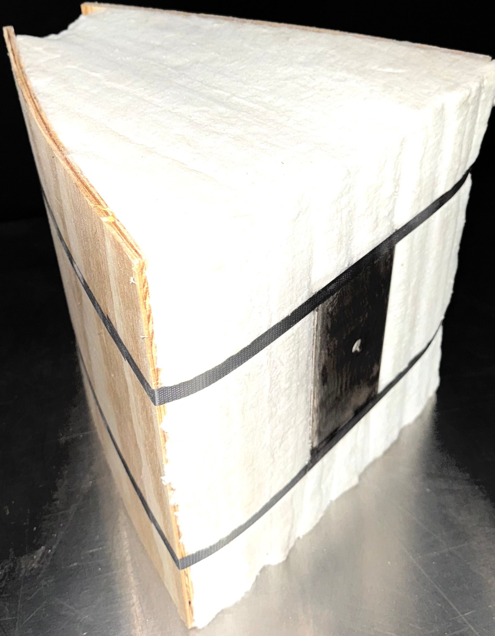 White, triangular packaged item held together with black straps. One side shows brown, layered material.