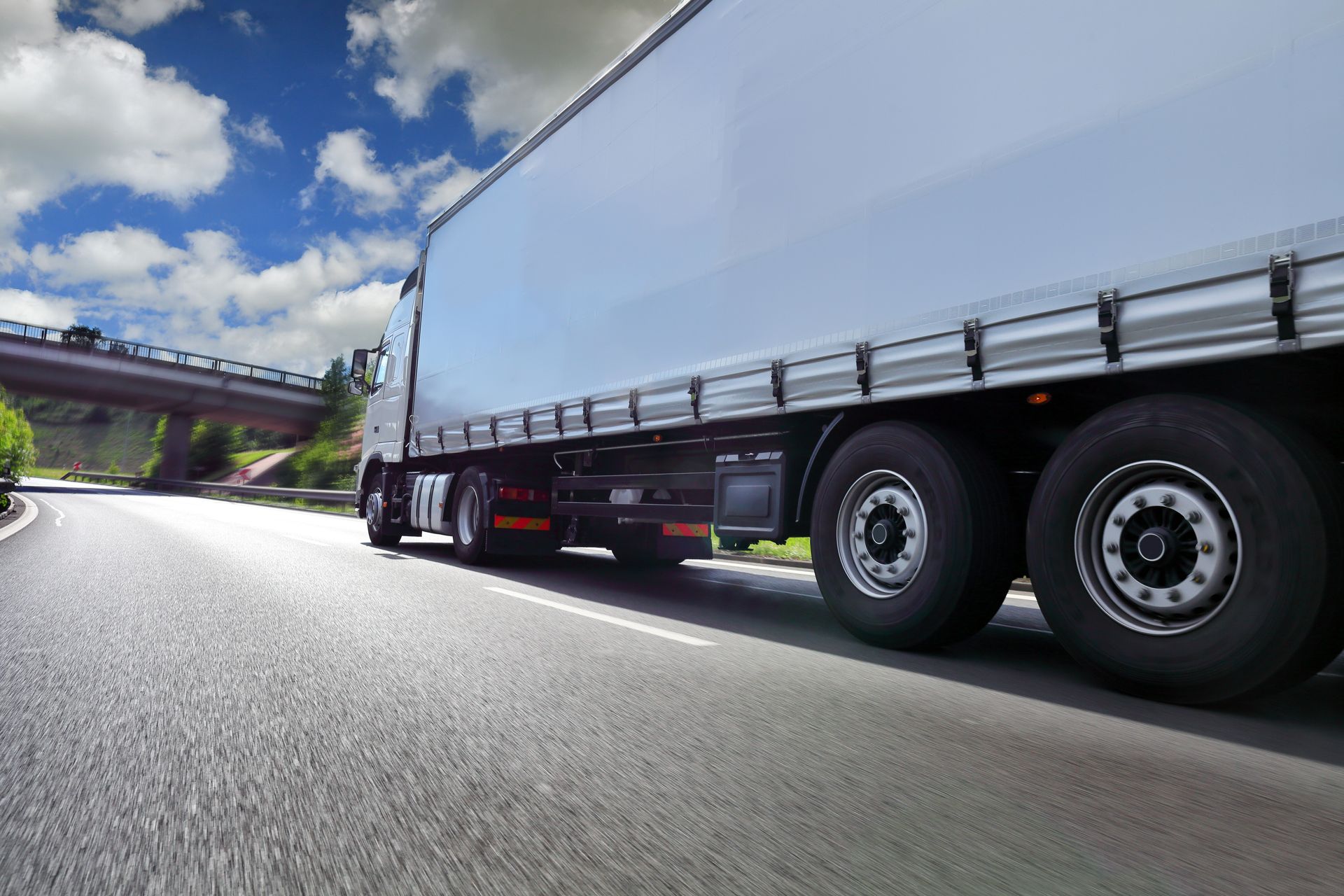 White semi-trailer truck driving on a highway under a bridge on a bright, cloudy day