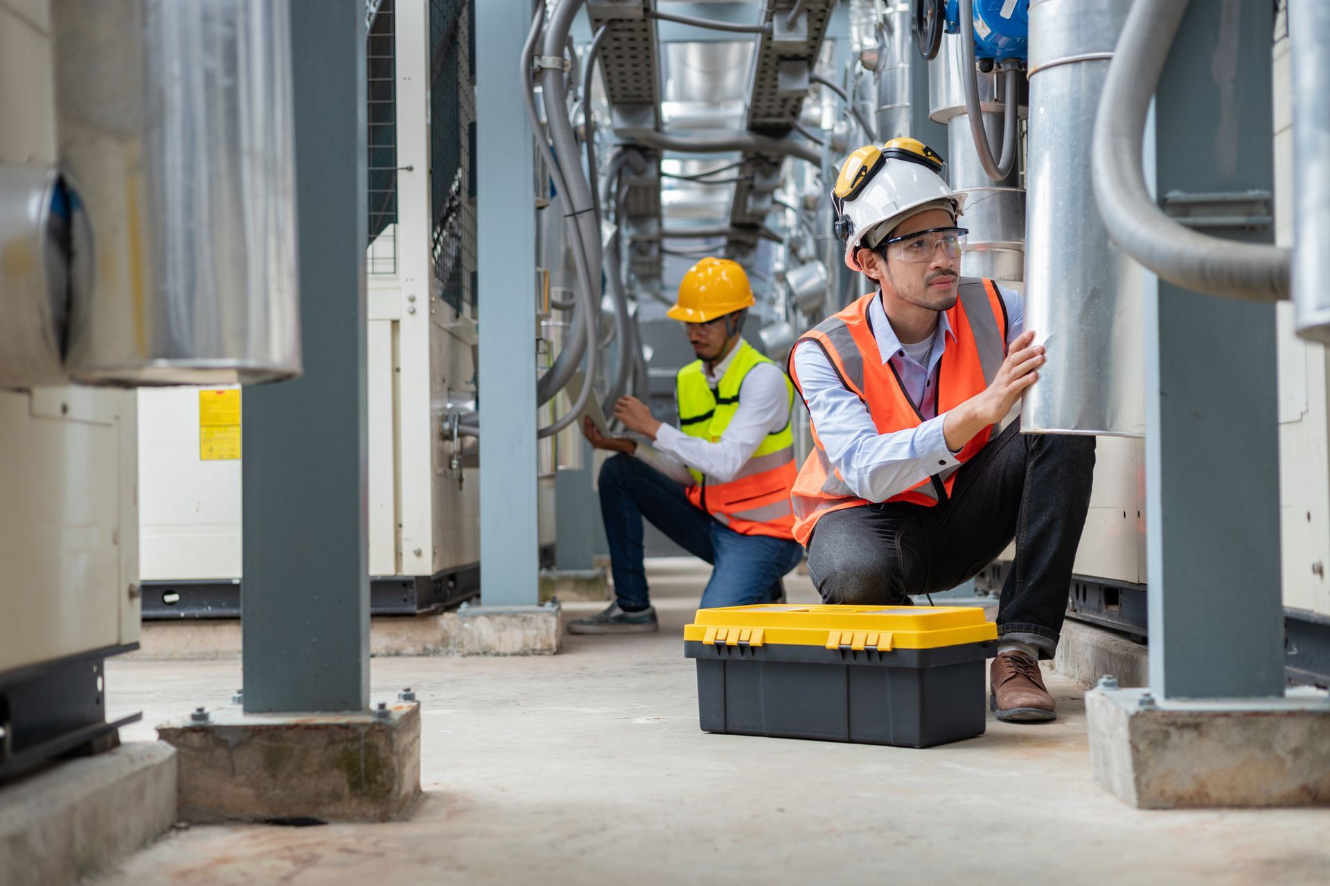 Two workers in safety gear inspect industrial equipment in mechanical room.