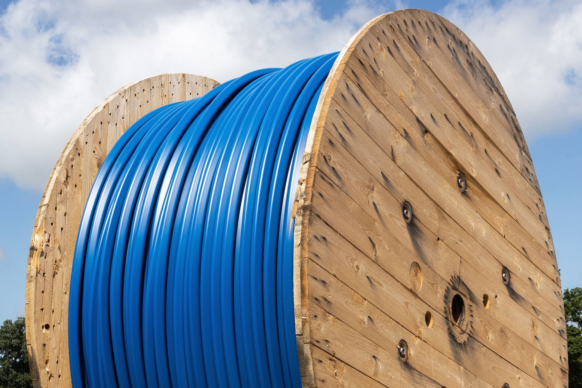 Large wooden spool holding blue ceramic fiber cable against sky background.
