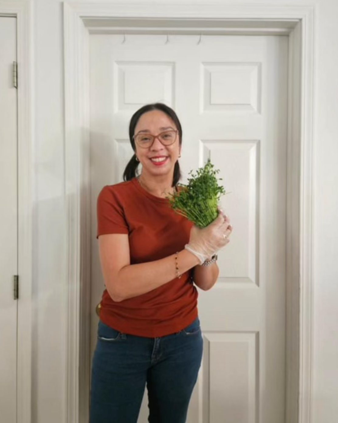 Woman in brown shirt and jeans, holding greens in front of a white door.