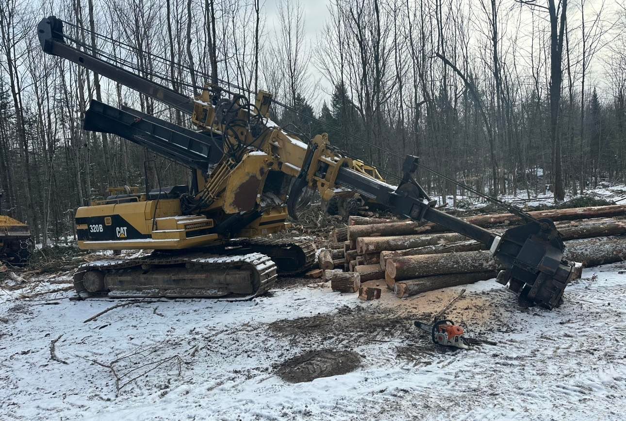 Une grande excavatrice jaune est assise dans la neige à côté d'un tas de bûches.