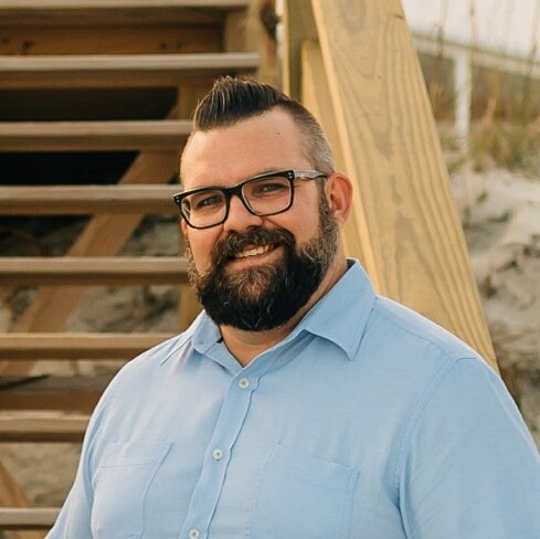 A man with a beard and glasses is standing in front of a wooden staircase.