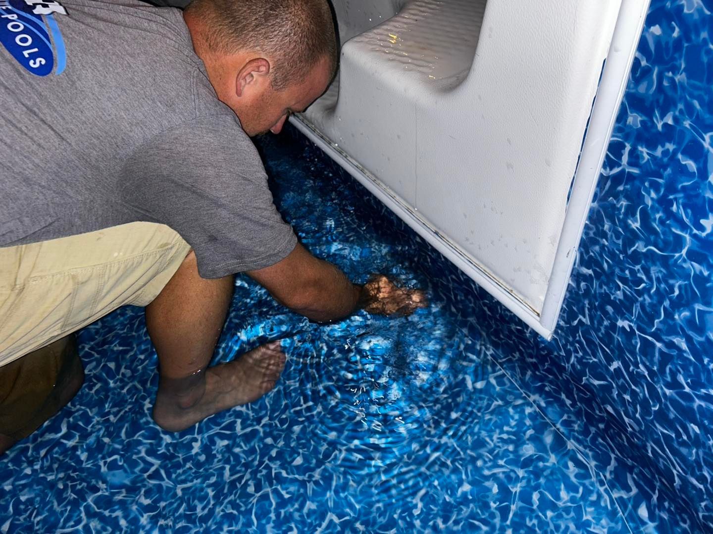 A service technician kneeling in a pool to repair the seam of a blue patterned vinyl liner at the base of stairs.