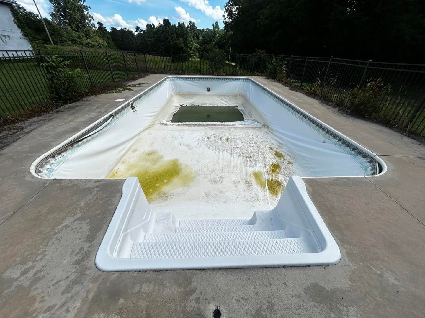 An empty, weathered backyard swimming pool with white steps and discolored, dirty liner in a fenced outdoor area.