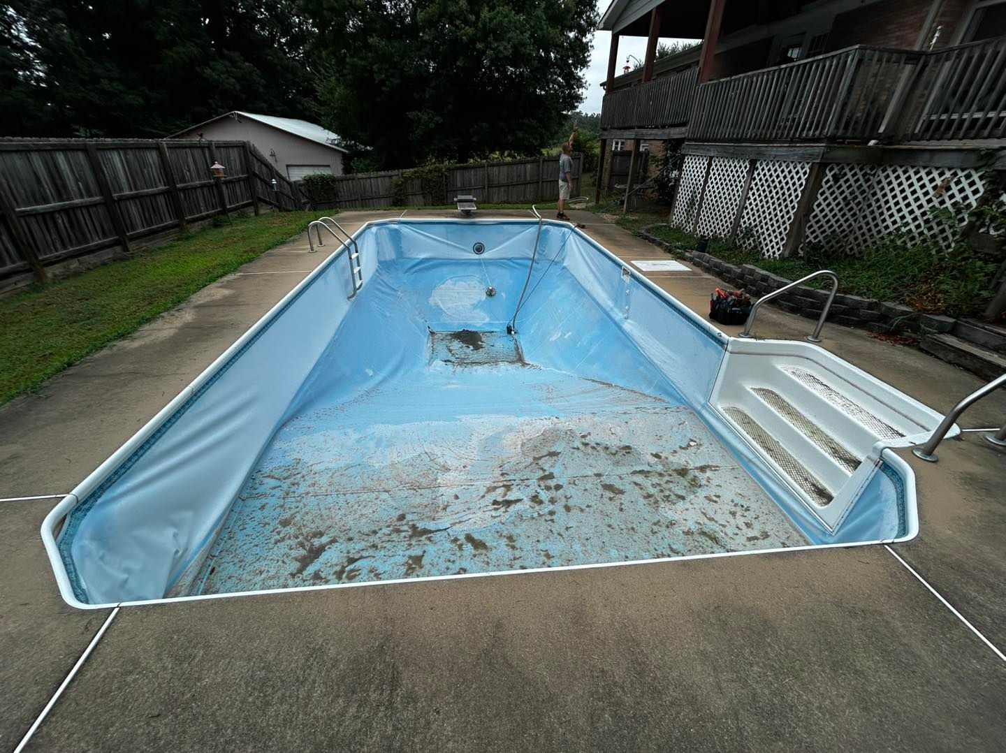 An empty, light blue rectangular swimming pool with debris on the floor and stairs on the right, set in a backyard.