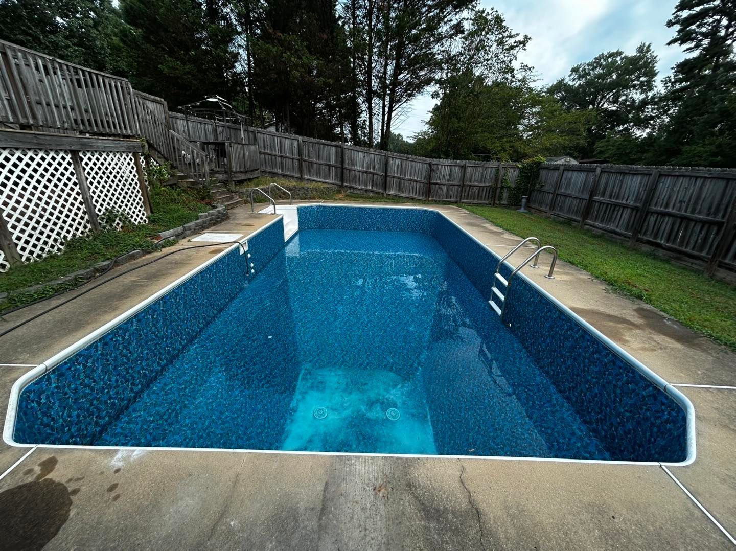 A rectangular, in-ground swimming pool with blue patterned lining, surrounded by a concrete deck and a wooden fence.