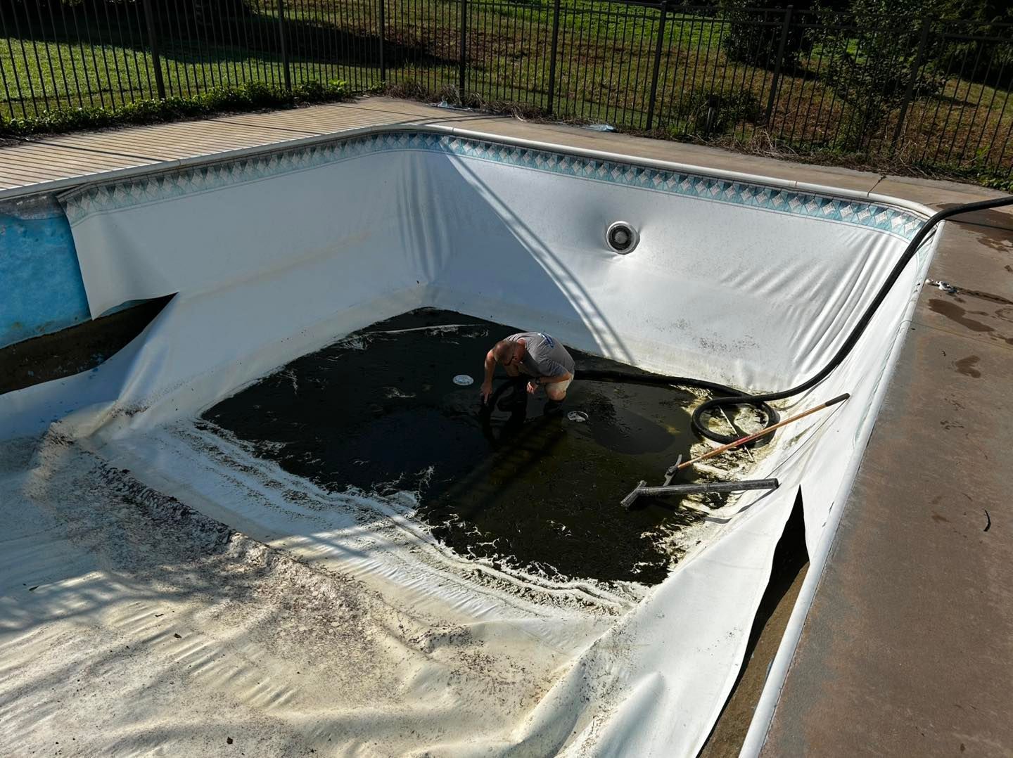 A person kneels in the center of an empty, white-lined swimming pool, working on the drain at the bottom.