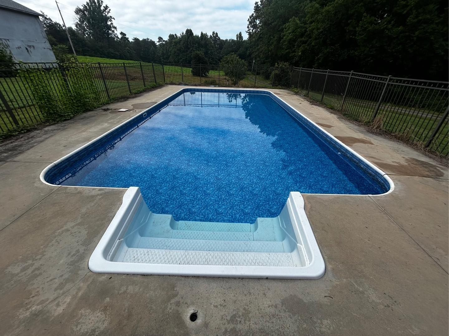A rectangular outdoor swimming pool with blue water and white entry steps, surrounded by a concrete deck and metal fence.