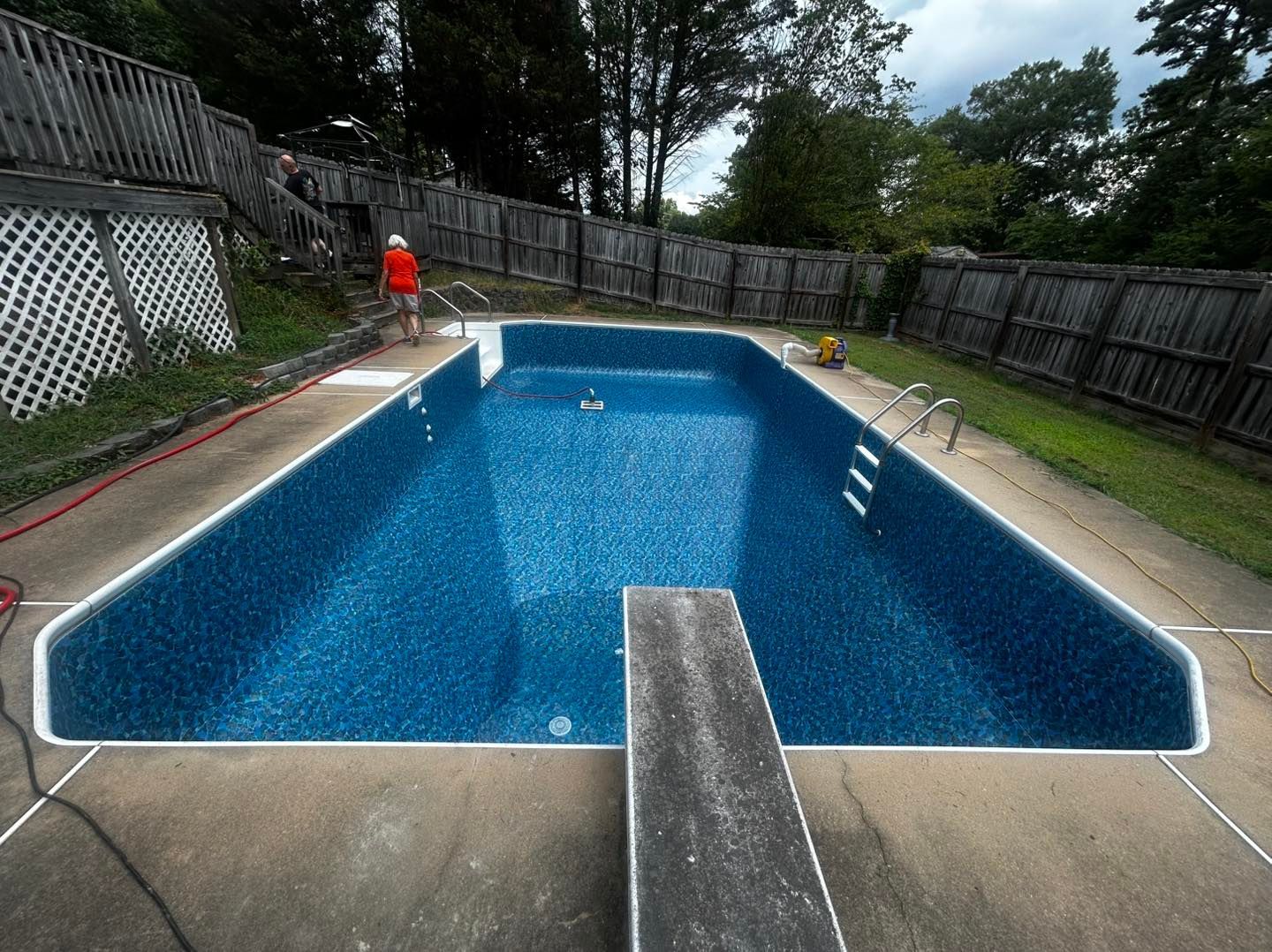 An empty rectangular swimming pool with blue patterned liner, a diving board, and a wooden fence in a backyard.