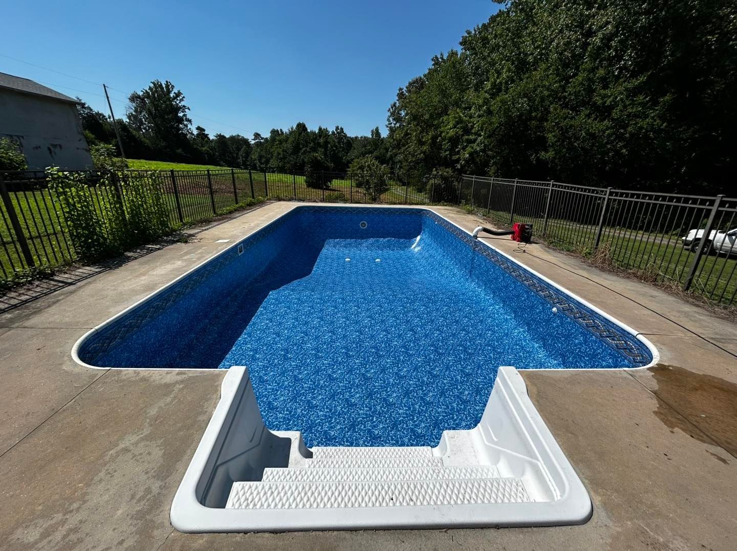 An empty backyard swimming pool with a white walk-in staircase, surrounded by a concrete deck and a fenced yard.