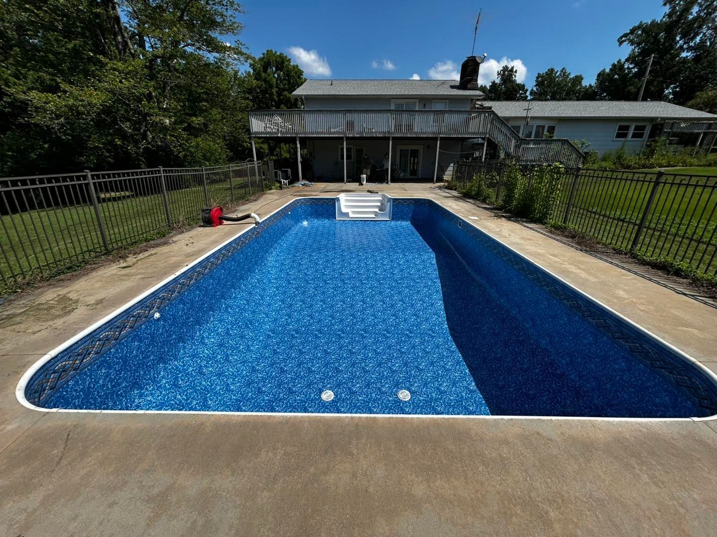 An empty rectangular swimming pool with blue patterned tile lining, set in a backyard near a house on a sunny day.