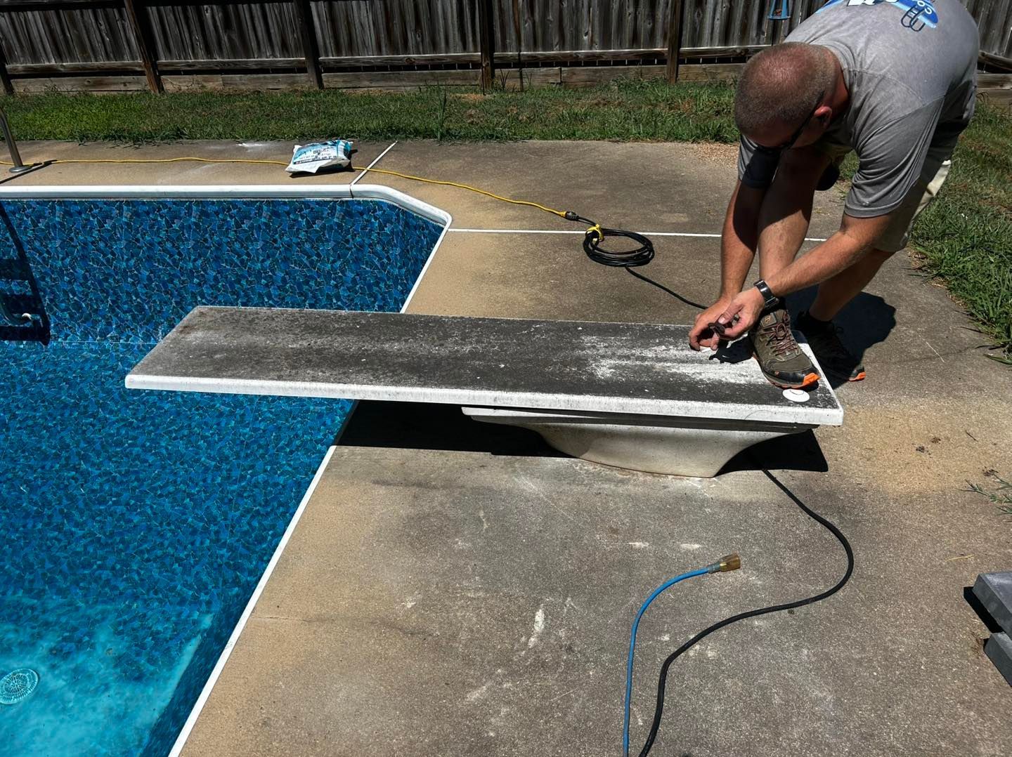 A person in a gray shirt kneeling on a concrete pool deck, working on a diving board near an outdoor swimming pool.