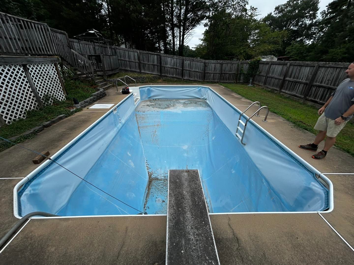 An empty, light-blue inground swimming pool with a diving board, set in a backyard with a person standing nearby.