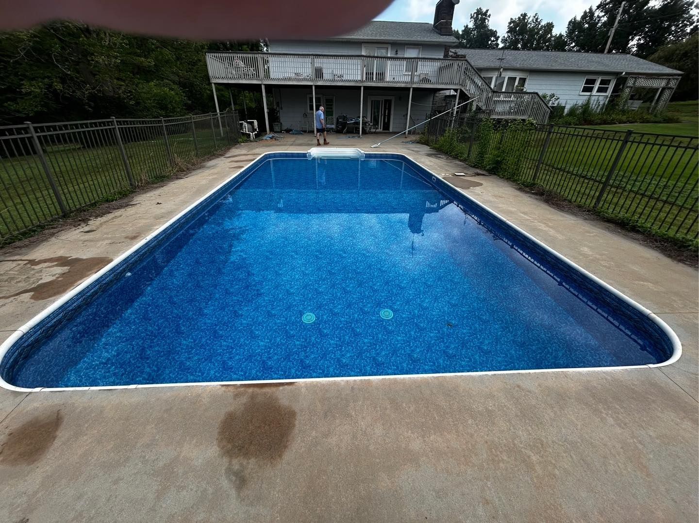 A rectangular, blue-lined in-ground swimming pool in a backyard, viewed from the concrete patio toward a house.