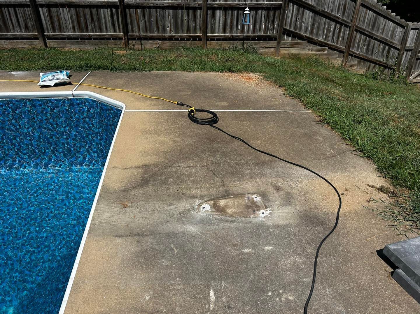 A black cord stretches across a stained concrete pool deck next to a blue tiled pool under a wooden fence.