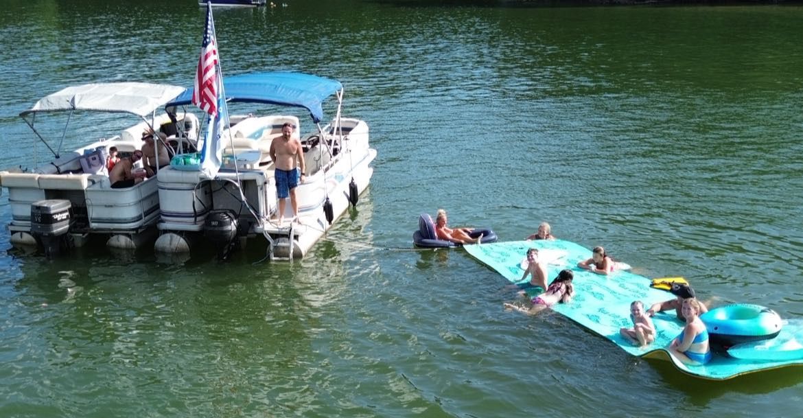 A group of people relaxing on a floating mat near two pontoon boats anchored in a lake.