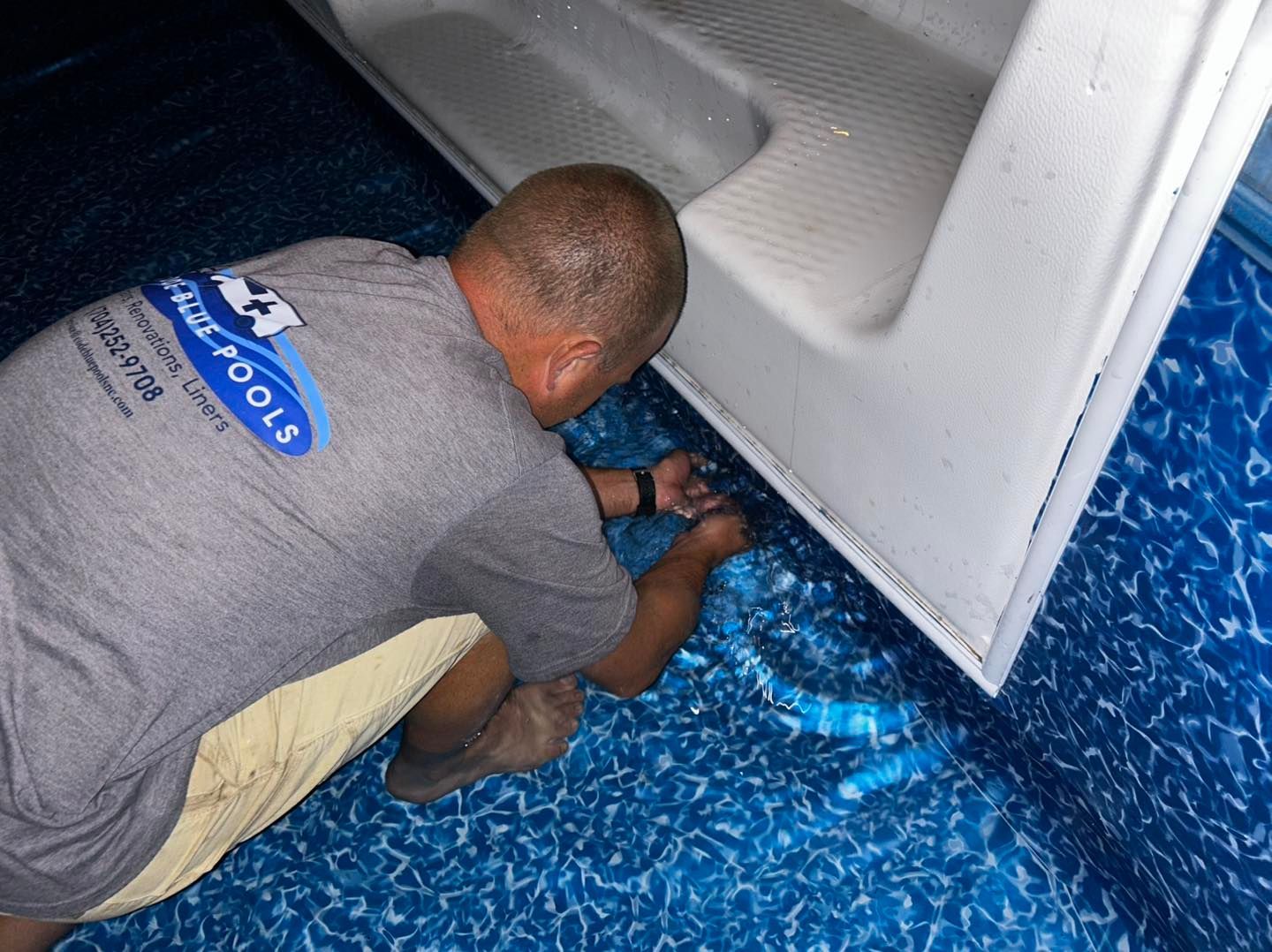 A person wearing a grey shirt works on the edge of a white pool step submerged in a blue patterned pool liner.