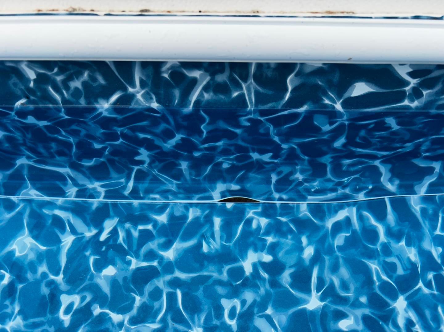 Close-up of a blue swimming pool liner with a visible horizontal tear or seam gap beneath the pool coping.