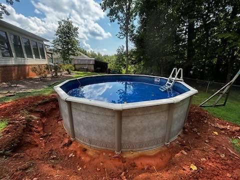A round, above-ground swimming pool filled with blue water, surrounded by red soil in a backyard near a house and trees.