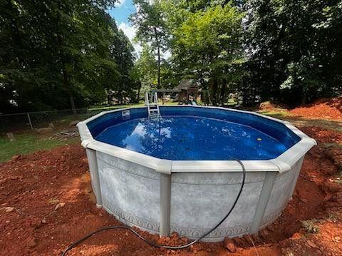 An above-ground, round pool with a grey exterior and blue water, situated in a dirt-filled yard surrounded by trees.