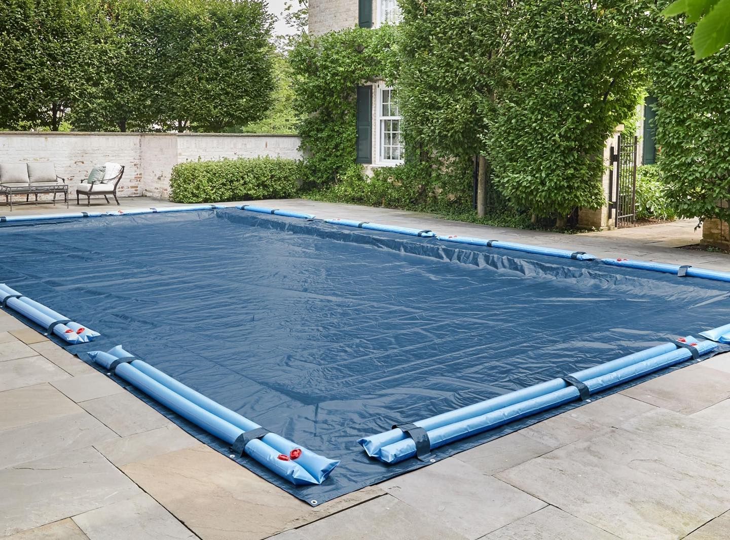 A rectangular swimming pool covered by a dark blue solar blanket, held in place by blue water-filled tubes on a stone patio.