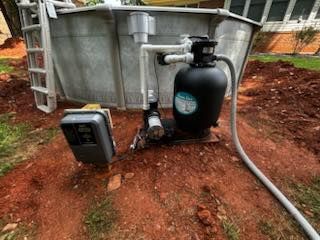 A swimming pool filter system and electrical box set in red dirt next to an above-ground pool.