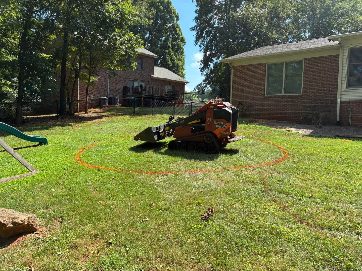 An orange skid steer sits in a residential backyard inside a circular marking sprayed on the grass.