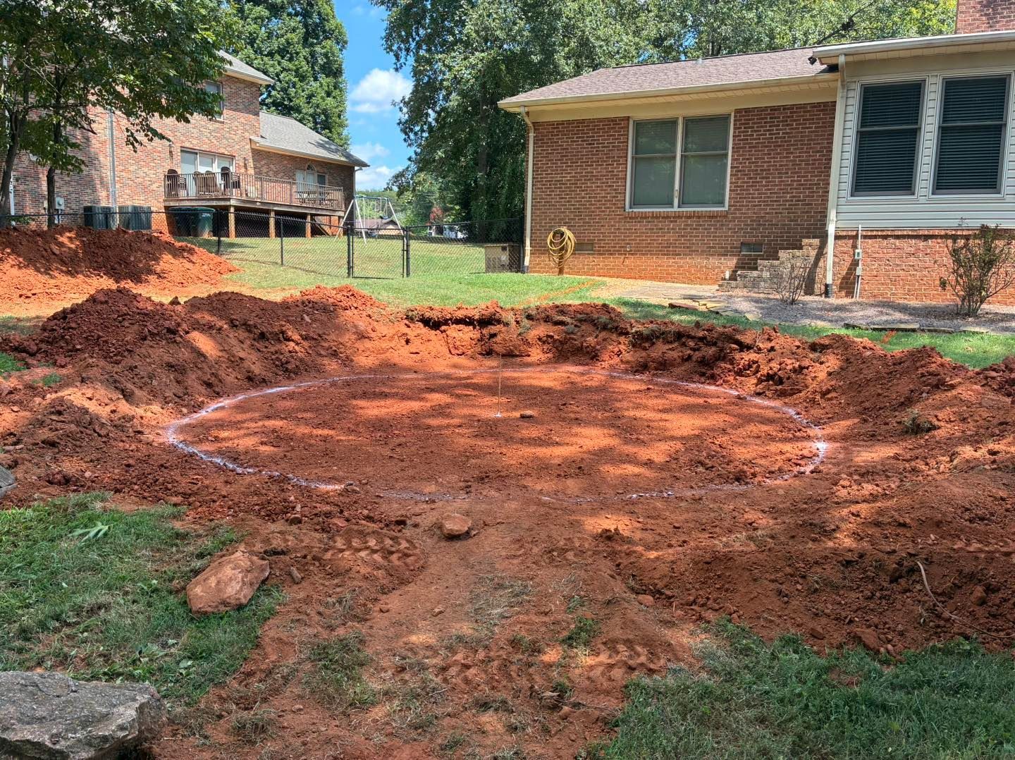 A circular area of excavated red clay soil sits in a residential backyard next to a brick house.