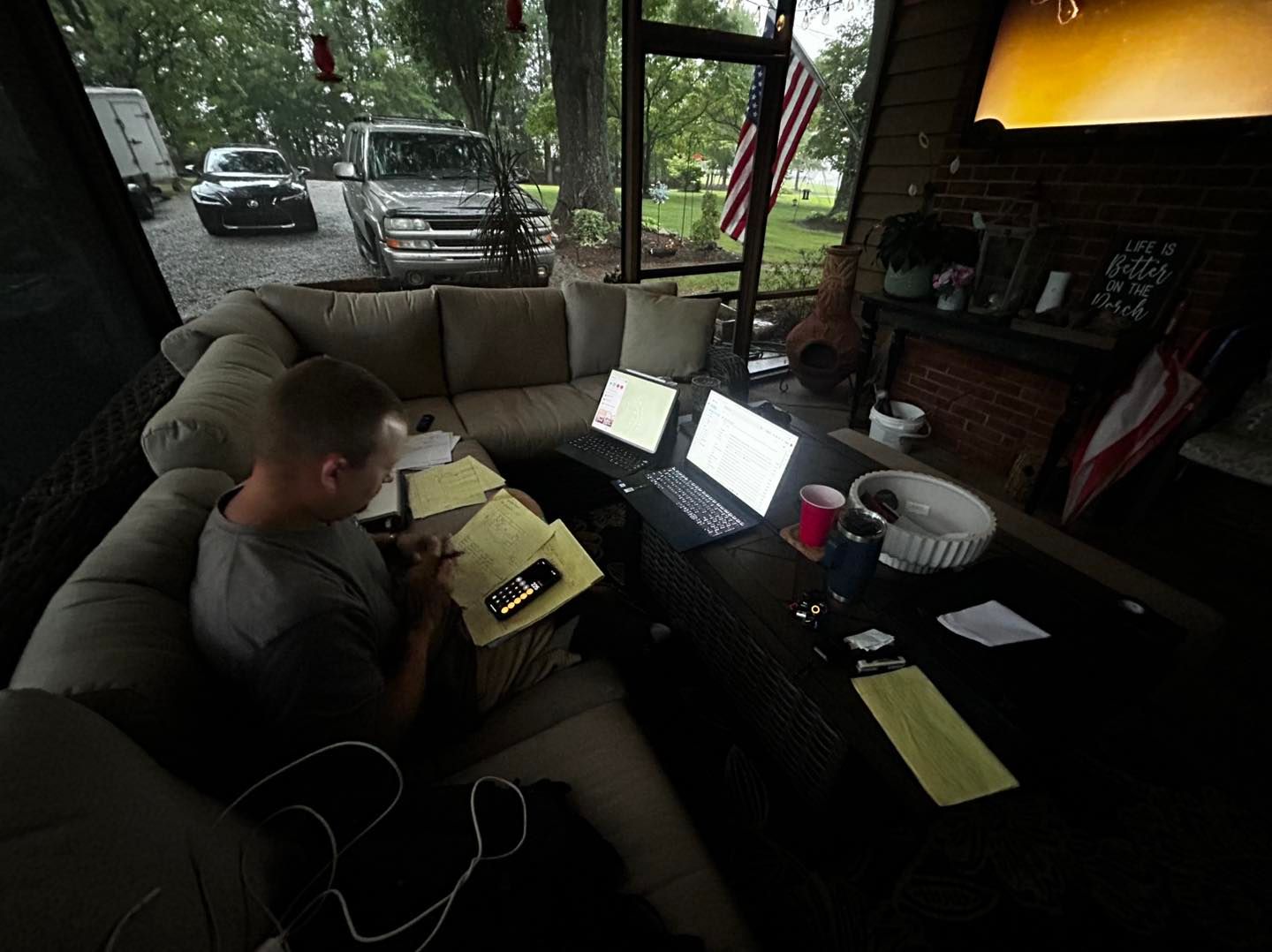 A person sits on a screened-in porch couch, working at a table with two open laptops, papers, and a smartphone.