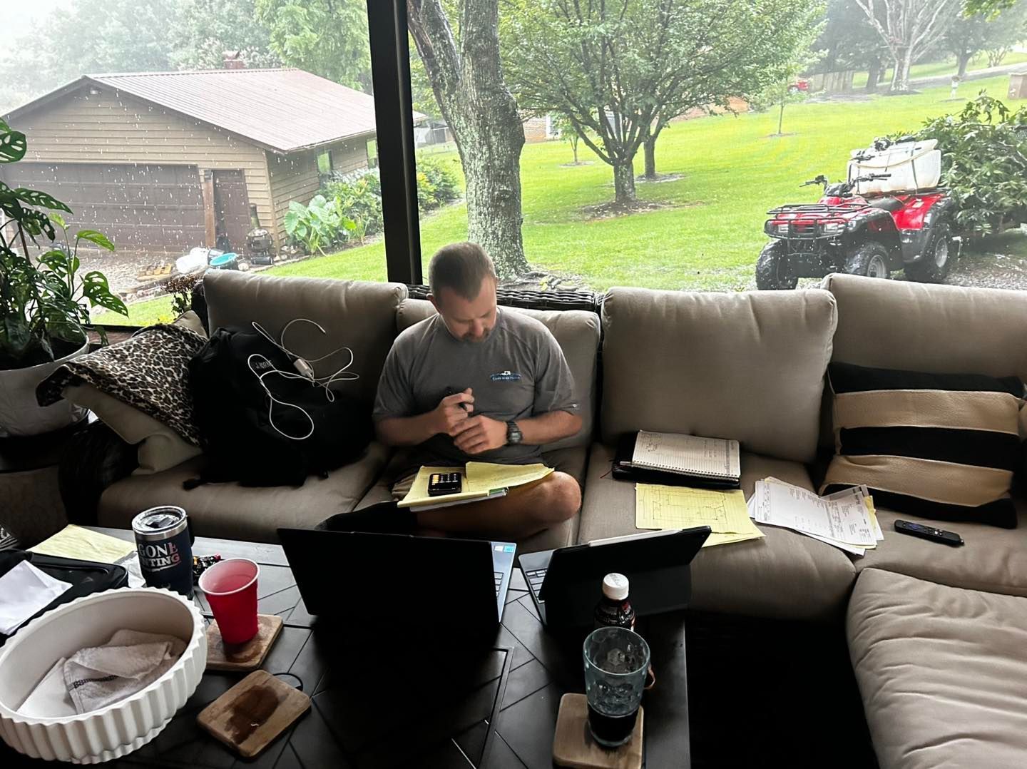 A person works on a laptop and documents while seated on a sectional sofa in a sunroom, with an ATV visible outside.