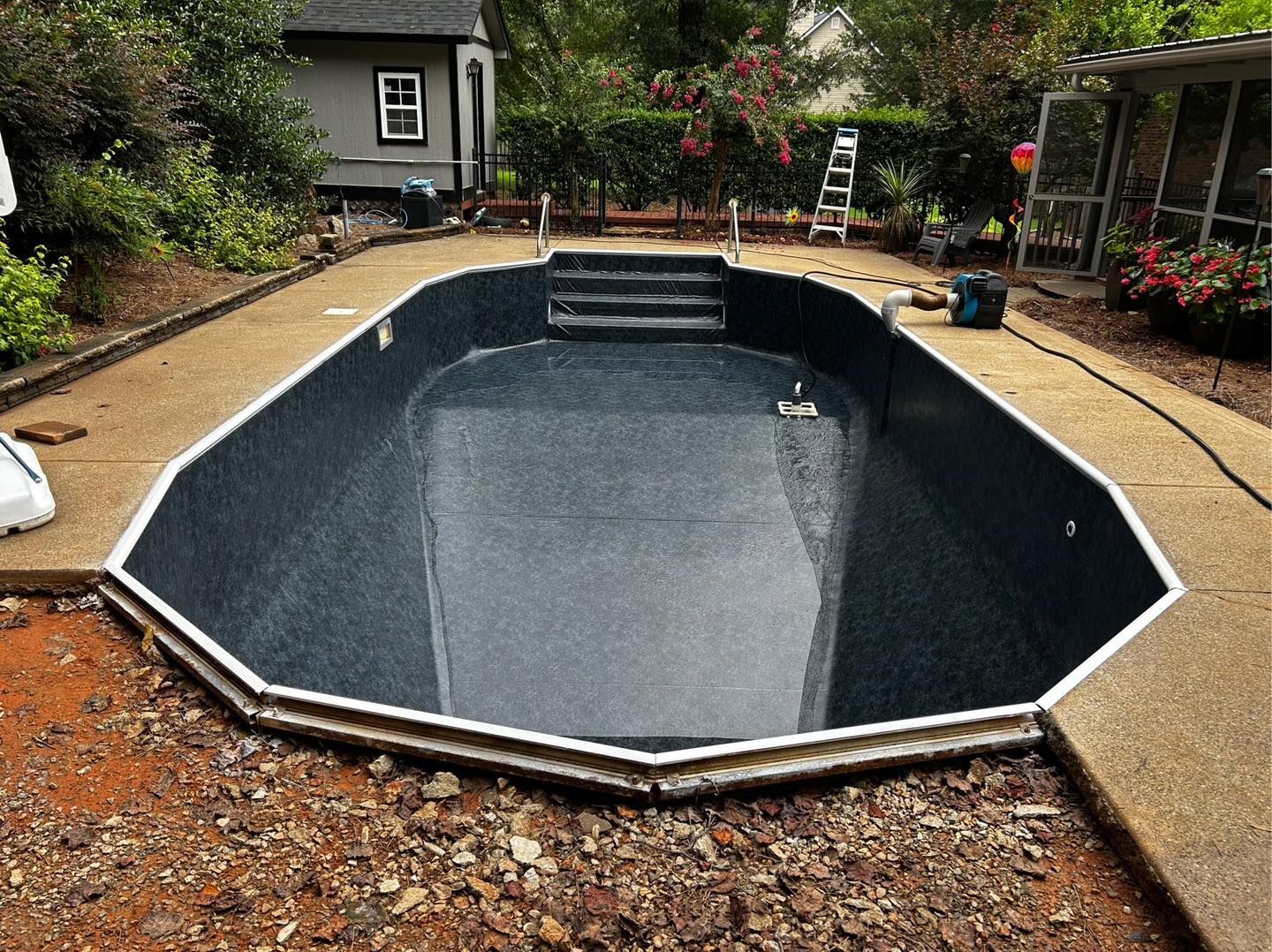An empty, dark-lined swimming pool with steps in a backyard setting under construction.
