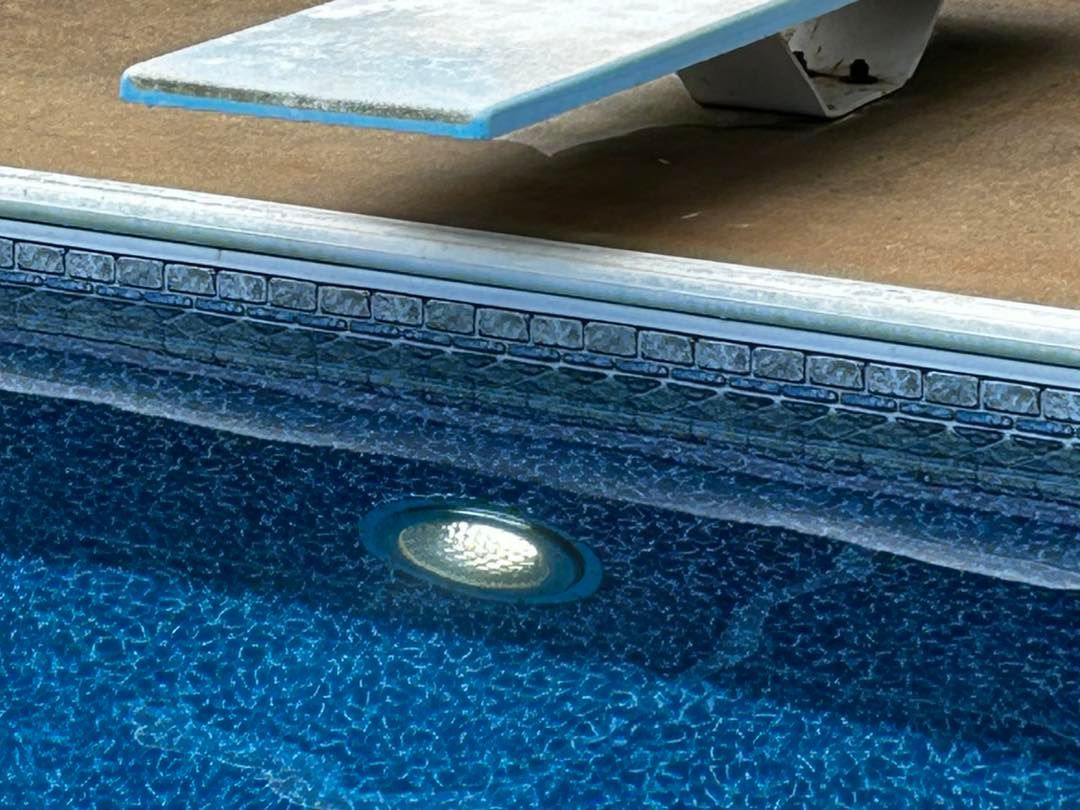 A close-up view of an underwater swimming pool light glowing against a patterned blue liner, below a white diving board.