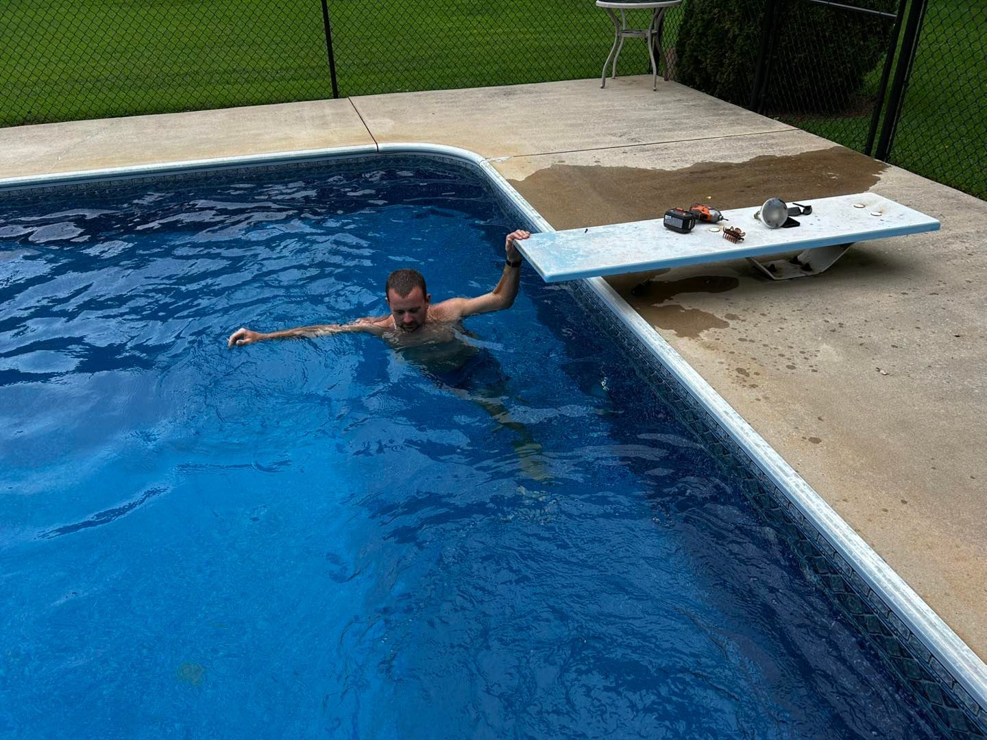 A person treads water in a blue swimming pool next to a diving board resting on the concrete deck.