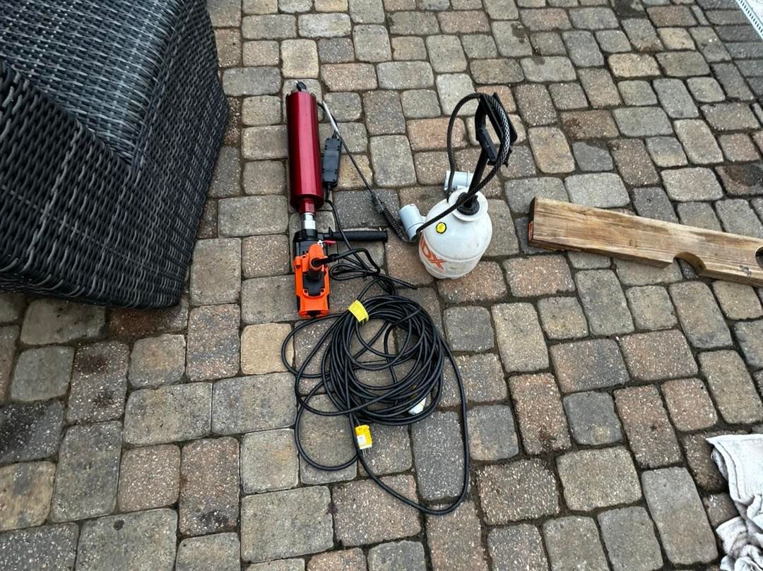 A diamond core drill, a water spray bottle, and a long power cord lying on a stone patio.