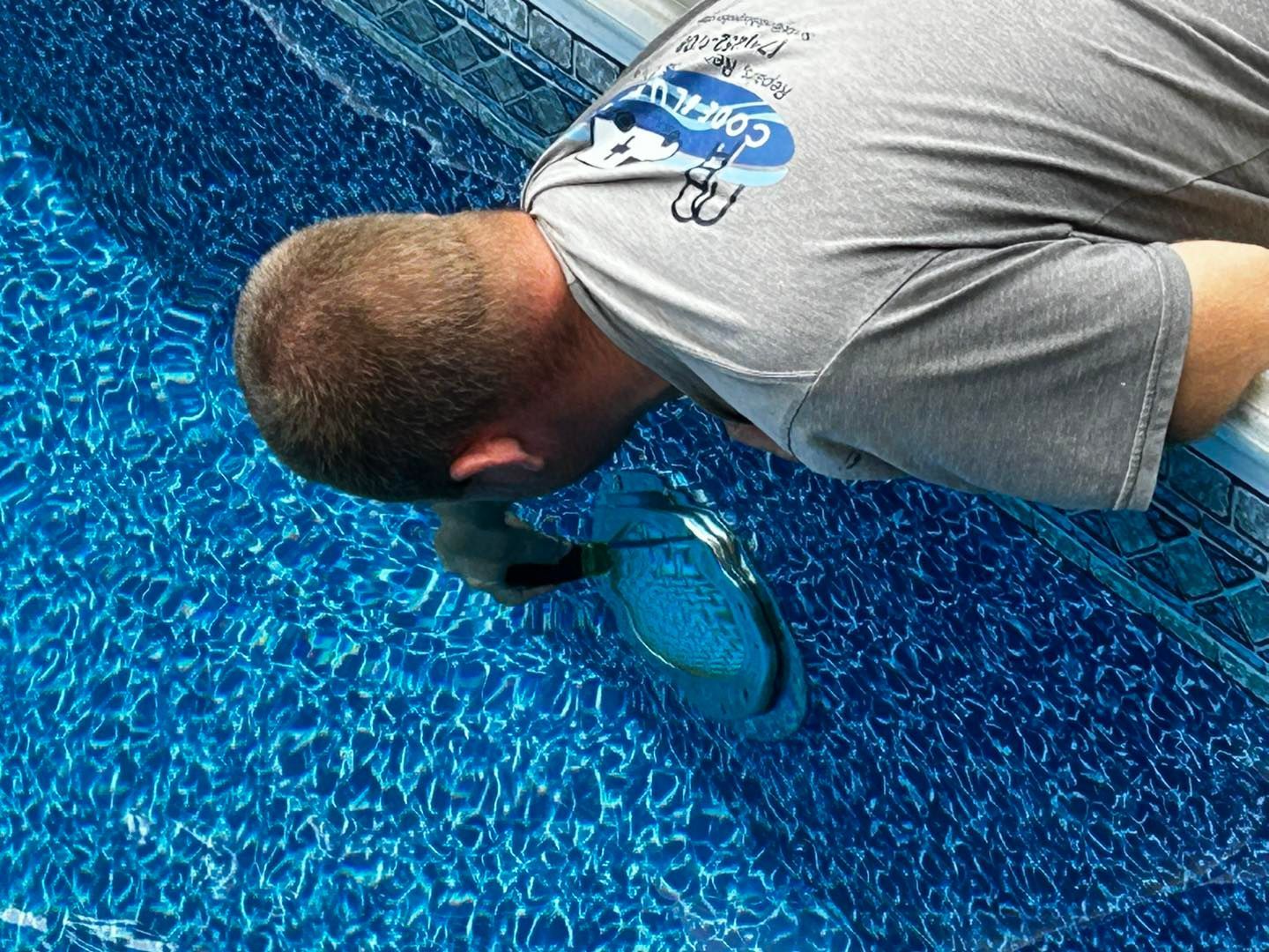 A person in a gray t-shirt leans over a pool edge, using a net to retrieve an object from the blue patterned water.