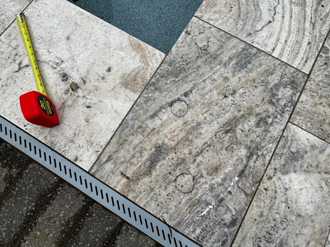 A close-up of a tape measure resting on light-colored stone pool deck tiles next to a linear drainage grate.