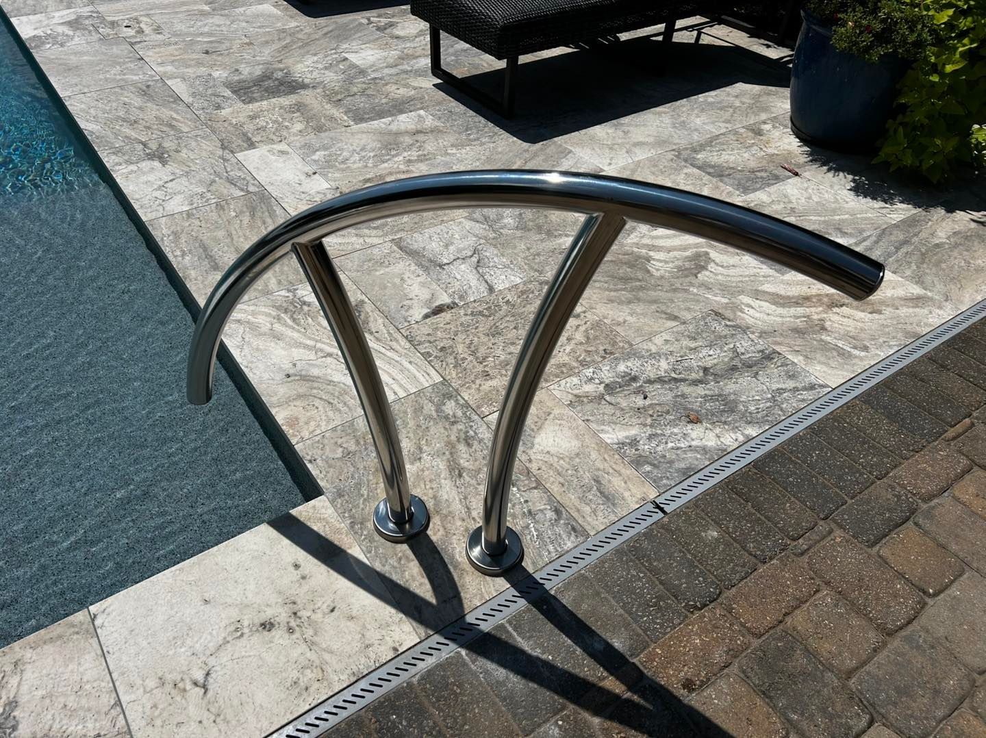 A polished stainless steel arched pool handrail mounted on stone patio tiles next to a swimming pool.