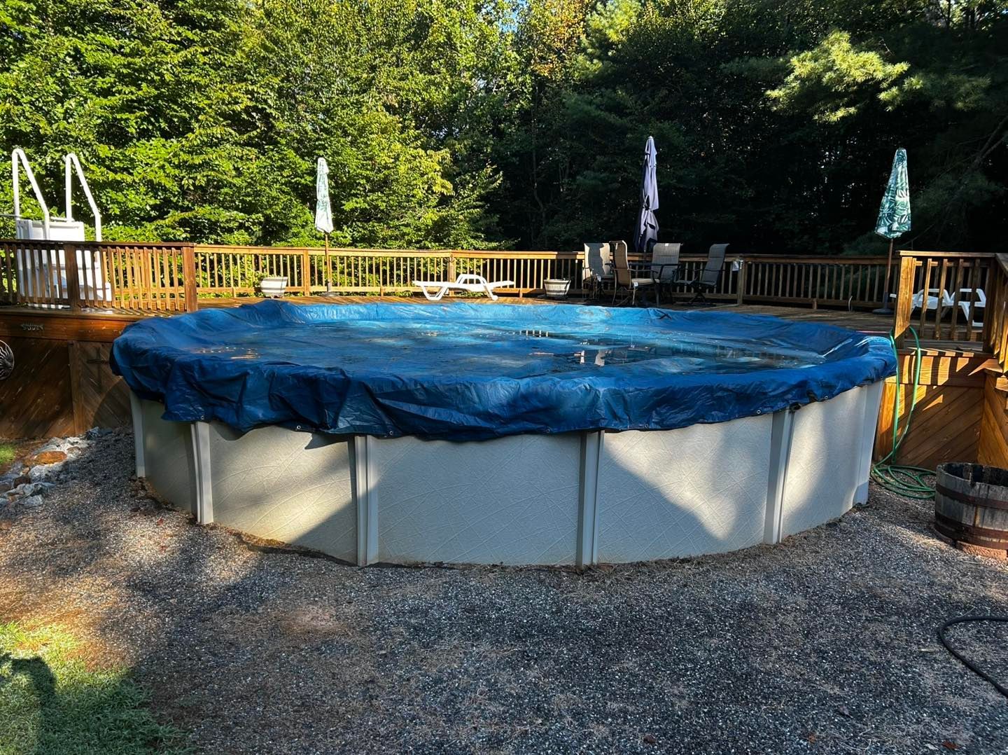 A round above-ground pool with a blue cover, set in a gravel yard next to a wooden deck surrounded by trees.