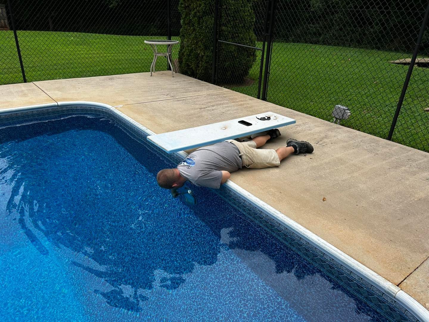 A person in a gray shirt and khaki shorts lies flat on a pool deck, looking down into the water near a diving board.