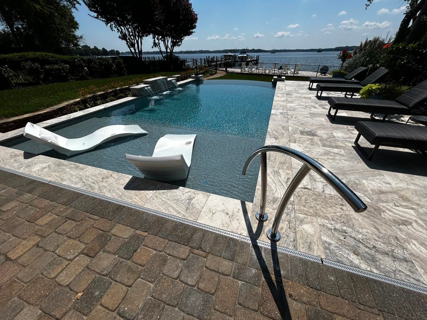 A rectangular swimming pool with two white lounge chairs on a shallow Baja shelf, overlooking a lake on a sunny day.