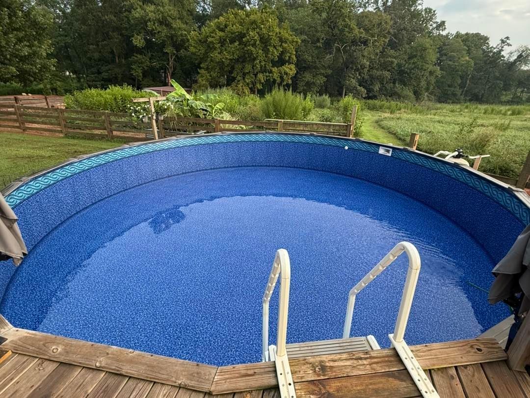 A round, above-ground swimming pool with blue patterned liner and white ladder, set in a grassy backyard near trees.