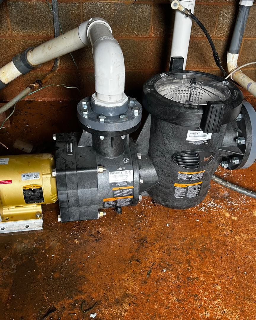 A yellow pool pump motor connected to a black plastic filter housing with white PVC piping against a brick wall.