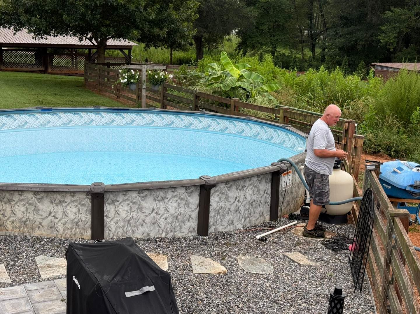 A person in a gray shirt working on a pool pump filter next to an above-ground pool in a backyard with a gravel patio.