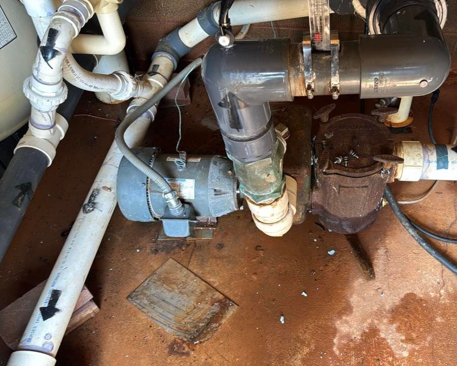 A pool pump system with grey and white PVC piping, a metal motor, and a rusty filter basket on a concrete surface.