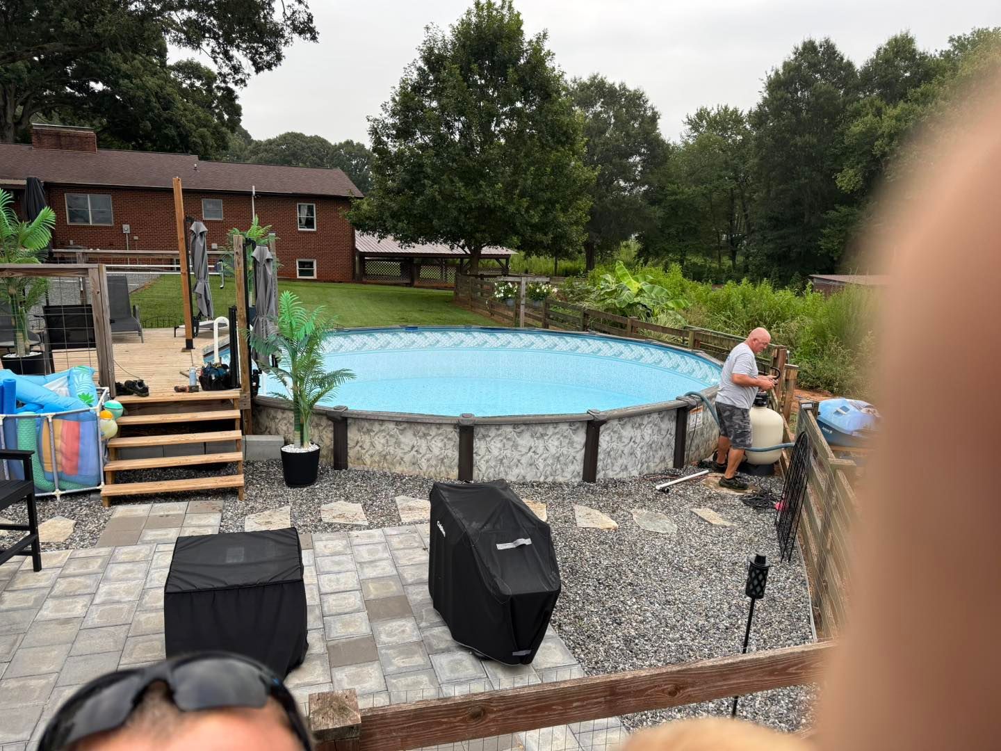 An above-ground pool in a backyard with a gravel patio, stone pavers, deck stairs, and a person standing by equipment.
