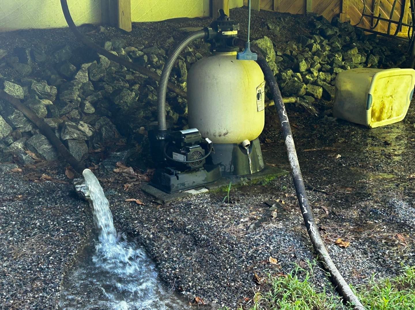 A pool pump and filtration tank sit on a gravel ground, with water gushing from a pipe into a small channel nearby.